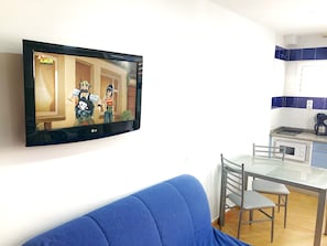 A flat-screen TV mounted on a wall, a blue sofa, a glass-top dining table with two chairs, and a kitchen area with blue and white tiles.