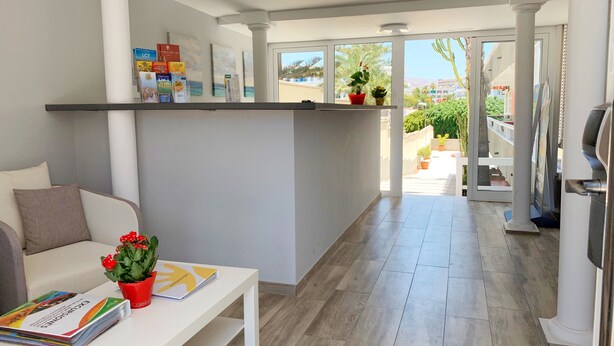 Una sala de estar moderna con un sofá blanco, una mesa de centro con revistas y una maceta roja, y un pasillo que conduce a un balcón con plantas en macetas.