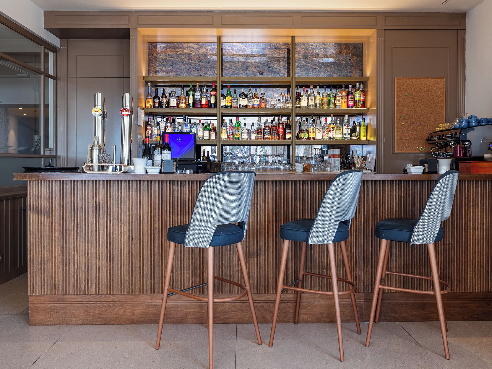 Een barruimte met een houten toonbank, meerdere flessenplanken, een digitaal scherm en drie barstoelen.