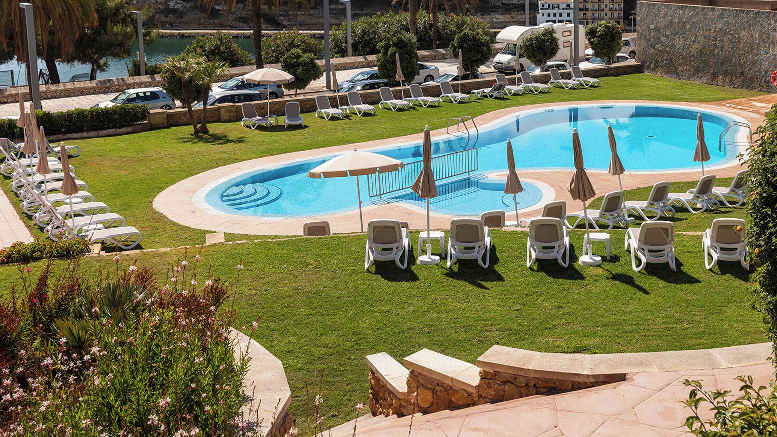 A swimming pool with lounge chairs and umbrellas, surrounded by a well-maintained lawn and a parking area with vehicles.