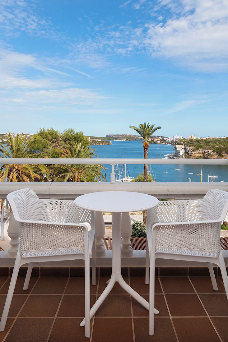 Une terrasse avec deux chaises blanches et une table, donnant sur une zone côtière où l’on aperçoit des bateaux et un ciel dégagé.
