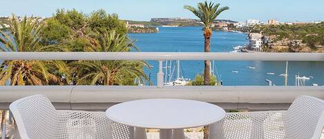 Une terrasse avec deux chaises blanches et une table, donnant sur une zone côtière où l’on aperçoit des bateaux et un ciel dégagé.