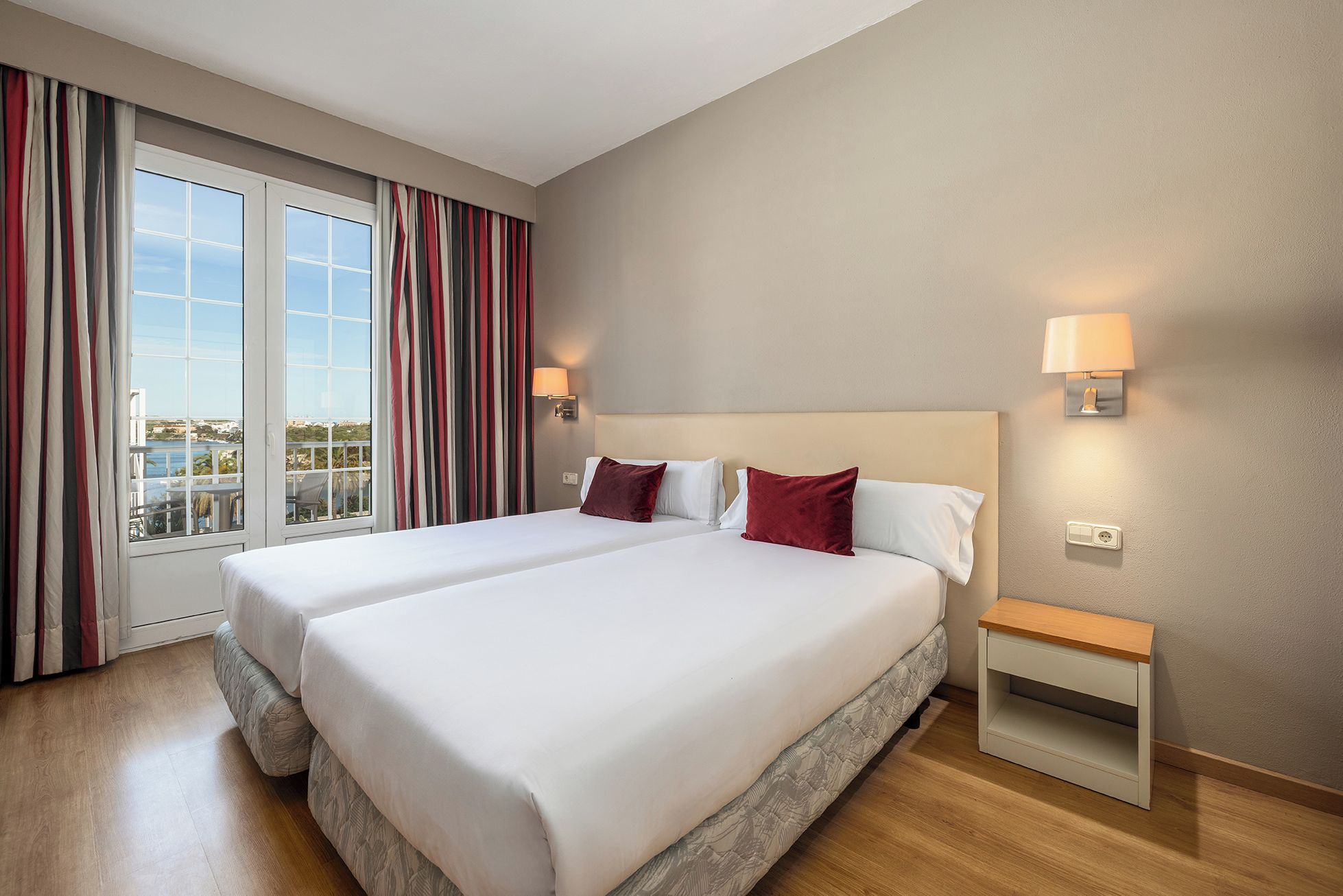 Ein Hotelzimmer mit zwei Betten, einem Balkon mit Vorhängen, einem Nachttisch und einer Wandlampe.