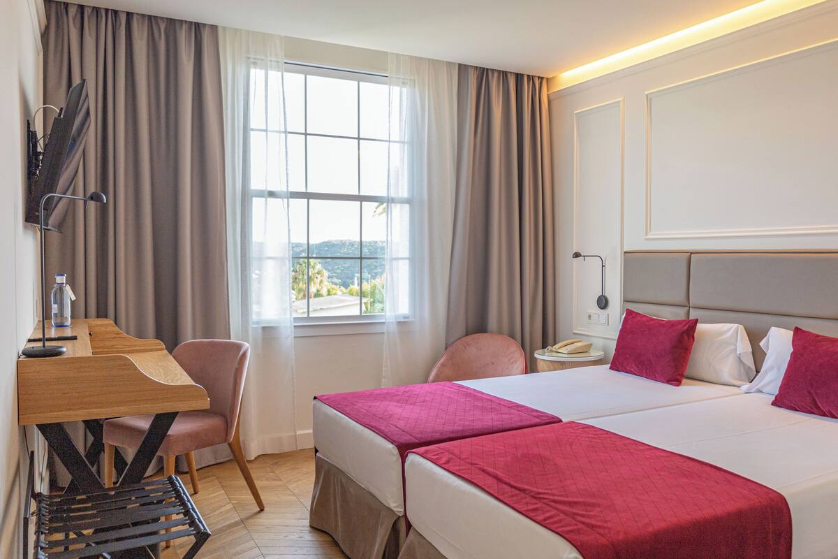 A hotel room with two beds, a desk, a chair, and a window with curtains.