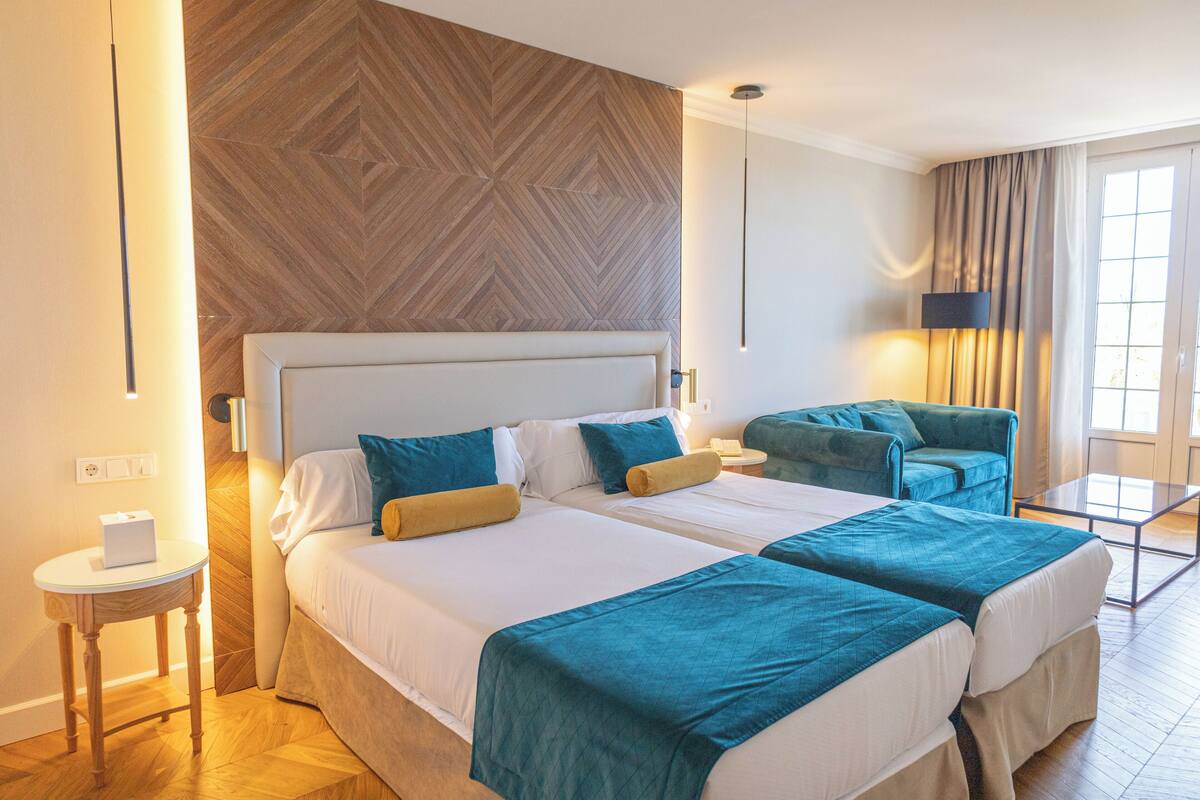 A hotel room with two beds, a wooden accent wall, a small table with a lamp, and a view of the outdoors through a window.