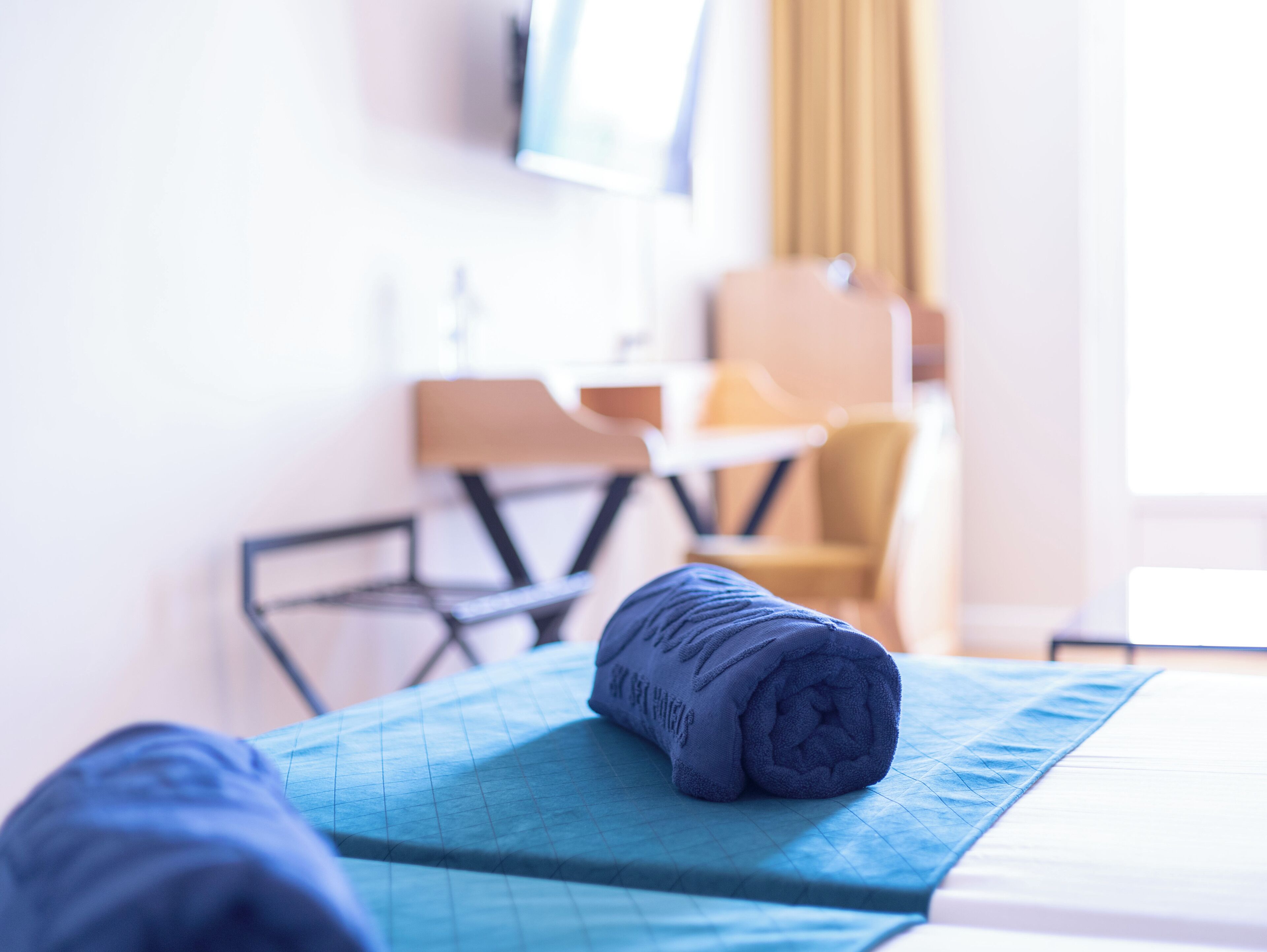 A neatly made bed with a blue blanket and a rolled-up towel on a blue mat.