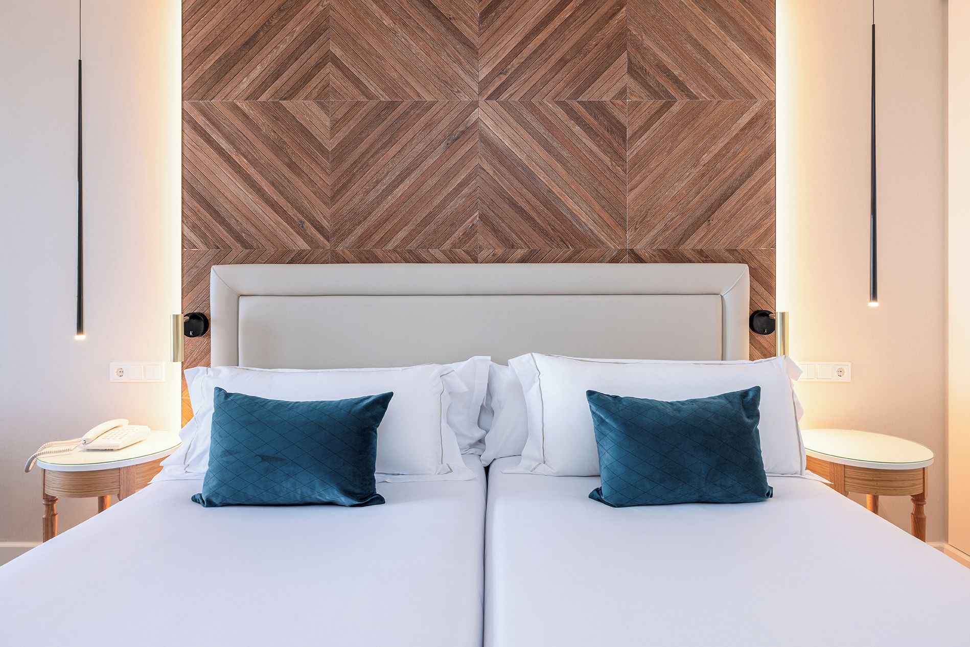 A double bed with white linens and two teal pillows, a wooden herringbone-patterned headboard, and bedside tables with lamps.