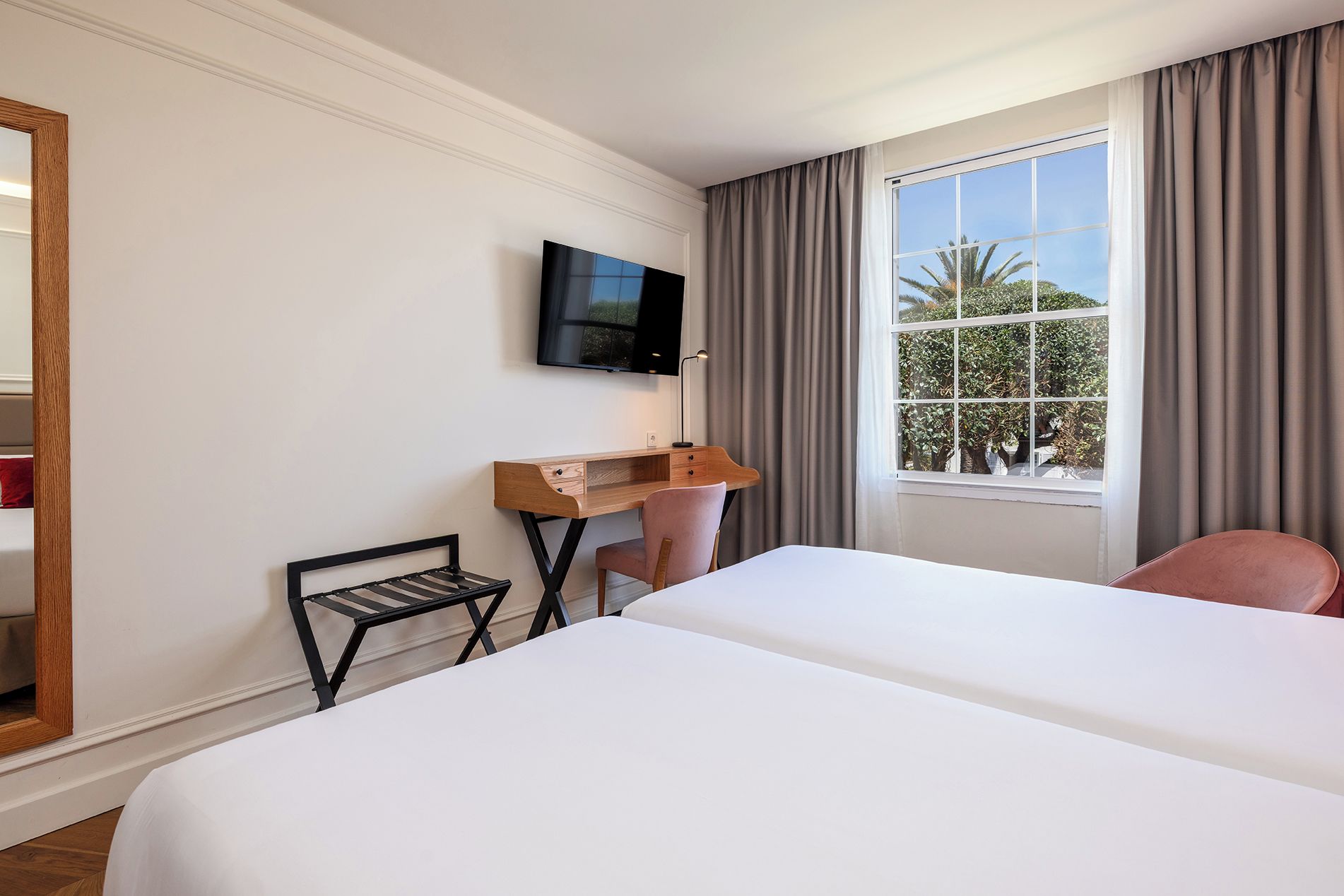 A hotel room with a large bed, a desk with a chair, a television mounted on the wall, and a window with curtains.
