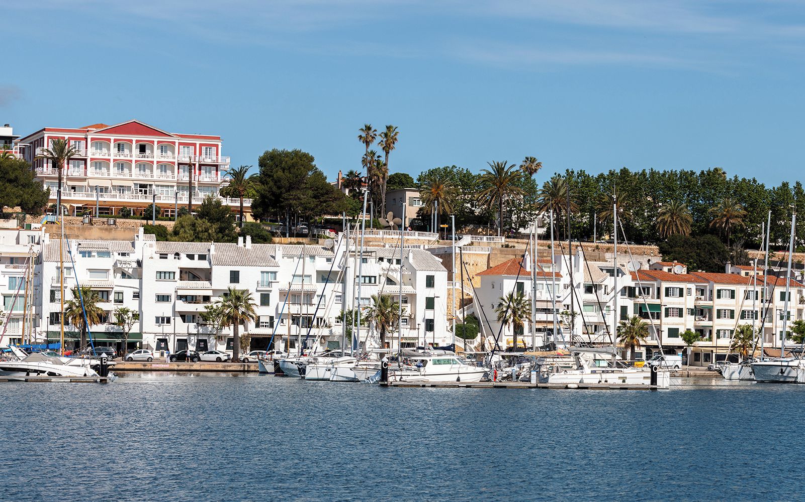A marina with multiple boats docked, surrounded by white buildings with balconies, and palm trees in the background.