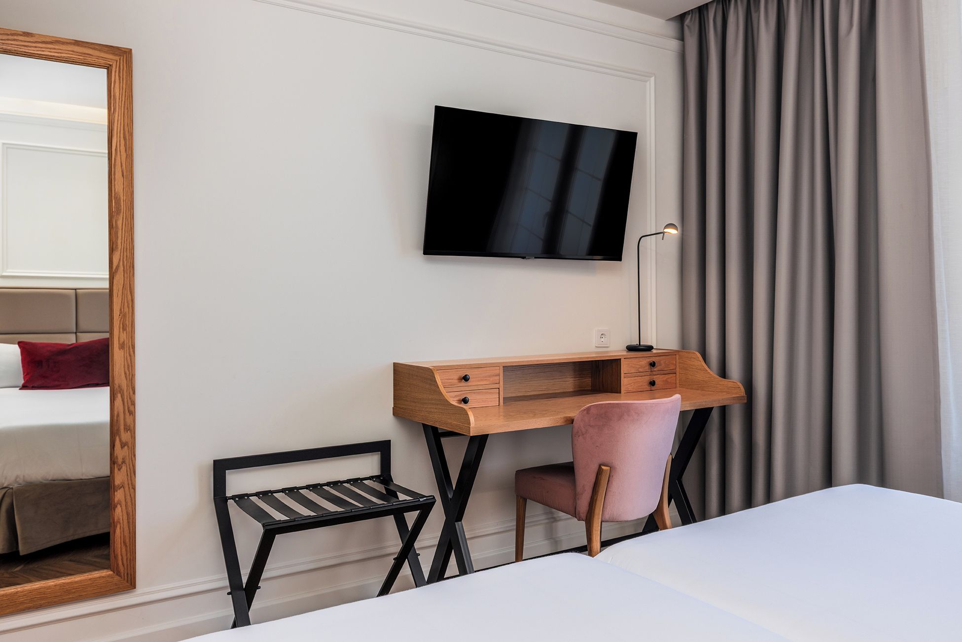 Habitación de hotel con cama, escritorio con silla, televisor montado en la pared y ventana con cortinas.