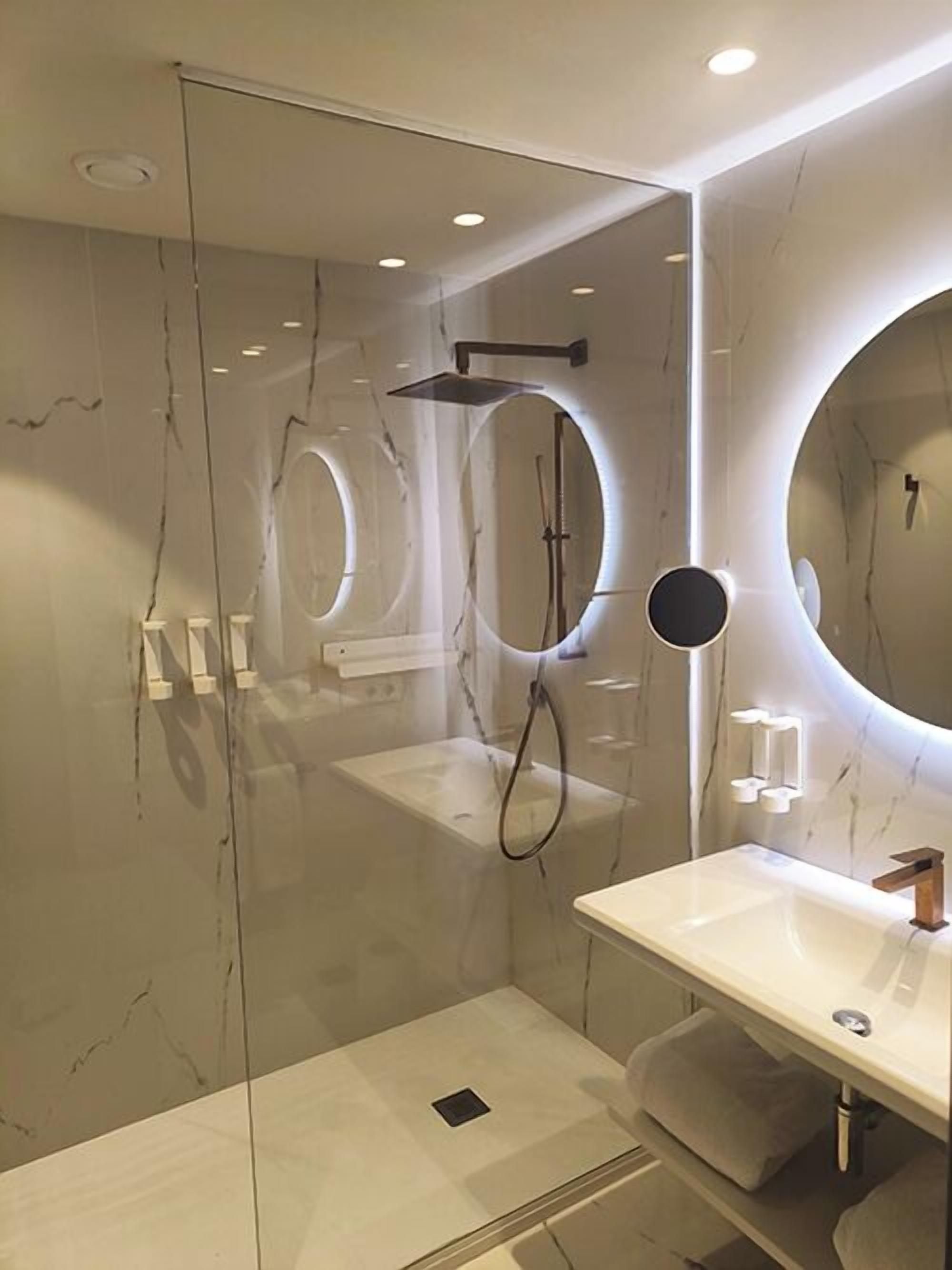 Une salle de bain moderne avec une douche à parois de verre, un miroir rond et un lavabo blanc.