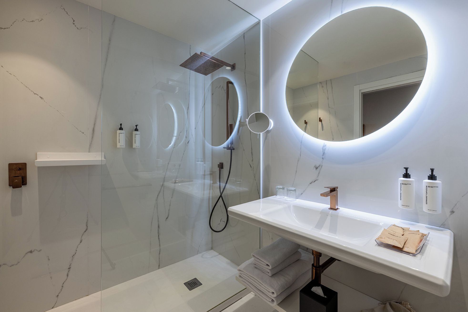 Une salle de bain moderne avec une douche à parois de verre, un grand miroir et un meuble-lavabo blanc où sont rangés les articles de toilette.