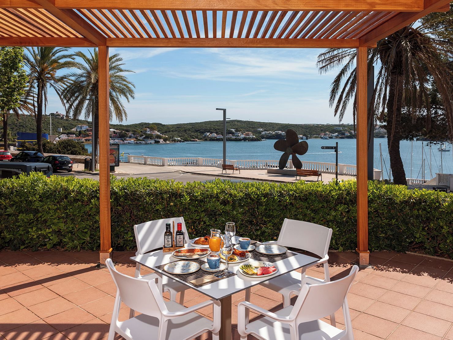 Un área de comedor al aire libre con una mesa para dos, con vista a la costa, una escultura y un puerto deportivo.