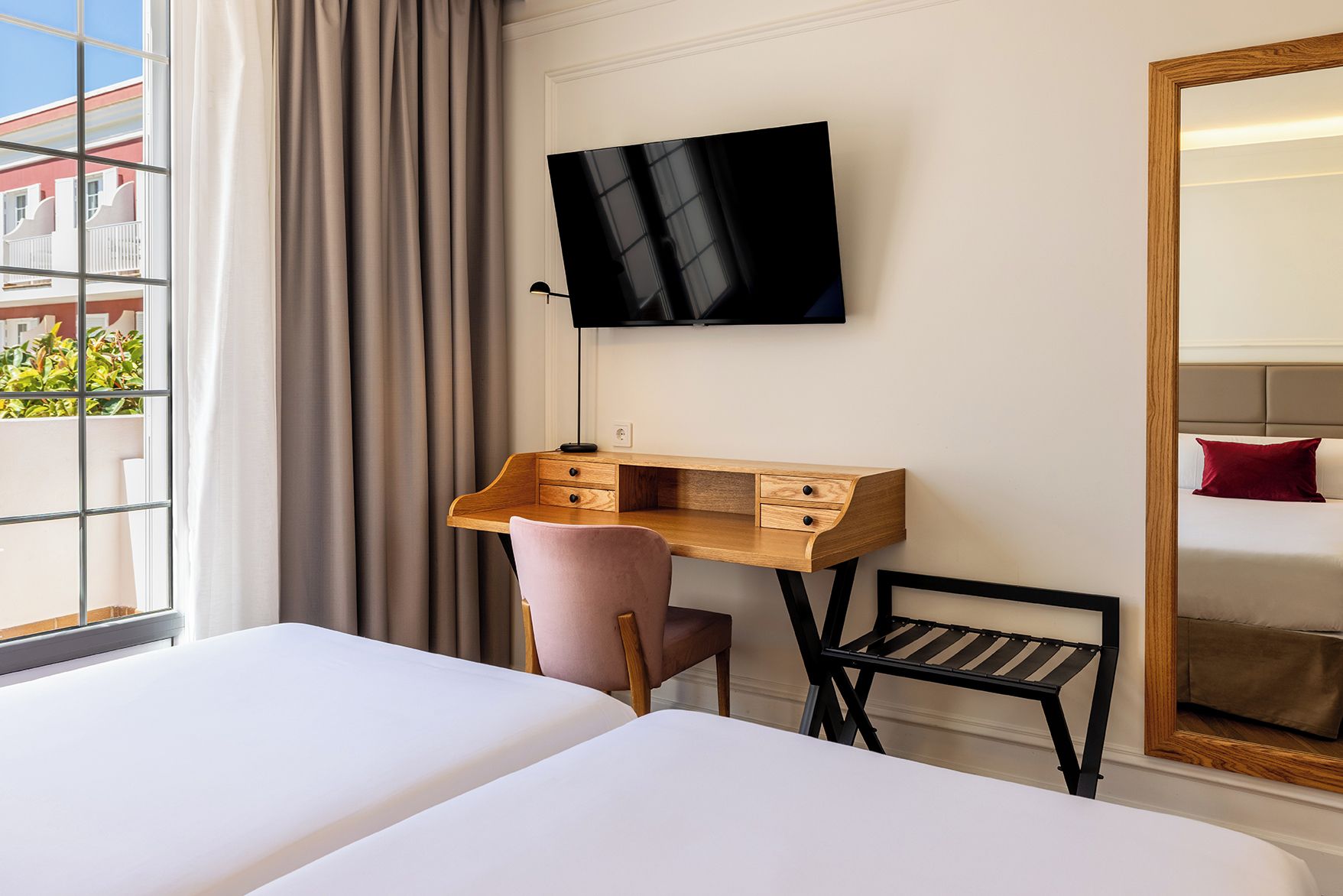 A hotel room with a bed, a desk with drawers, a chair, a TV mounted on the wall, and a large mirror.