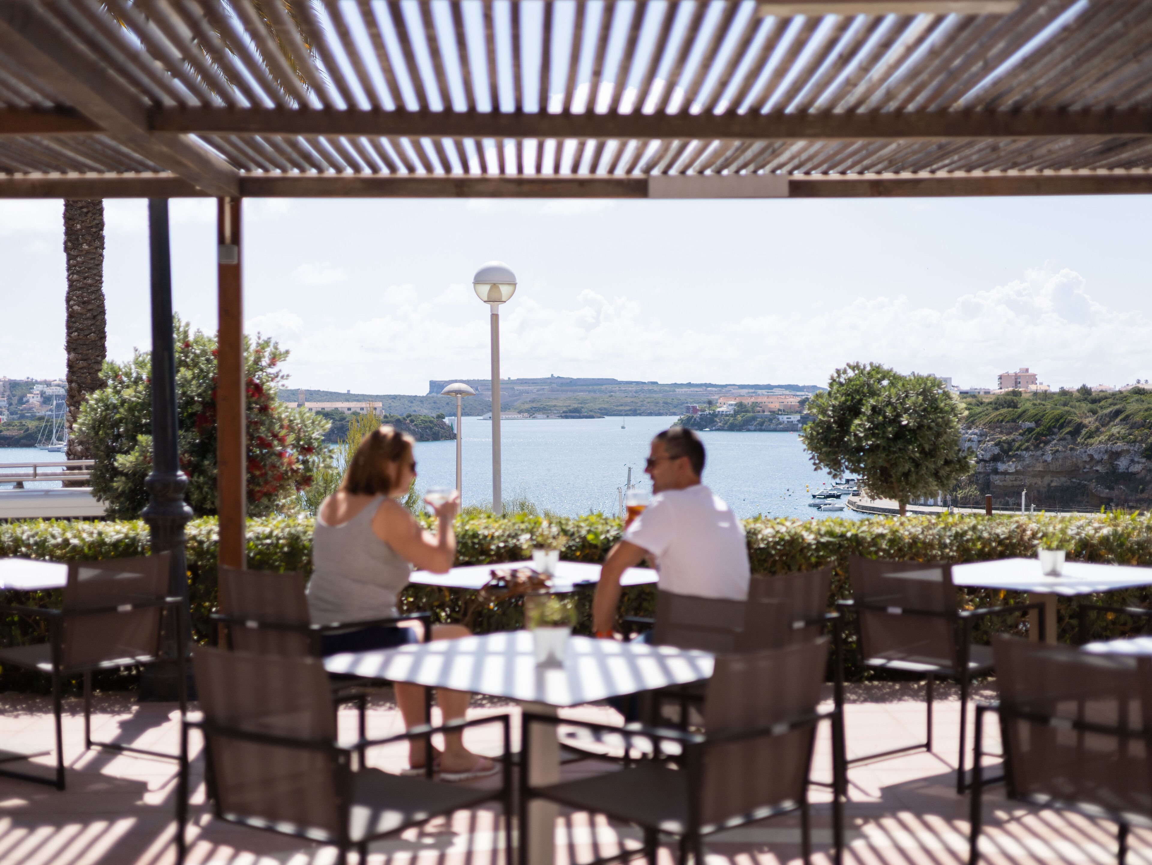 An outdoor patio with tables and chairs, overlooking a body of water and a distant landscape.