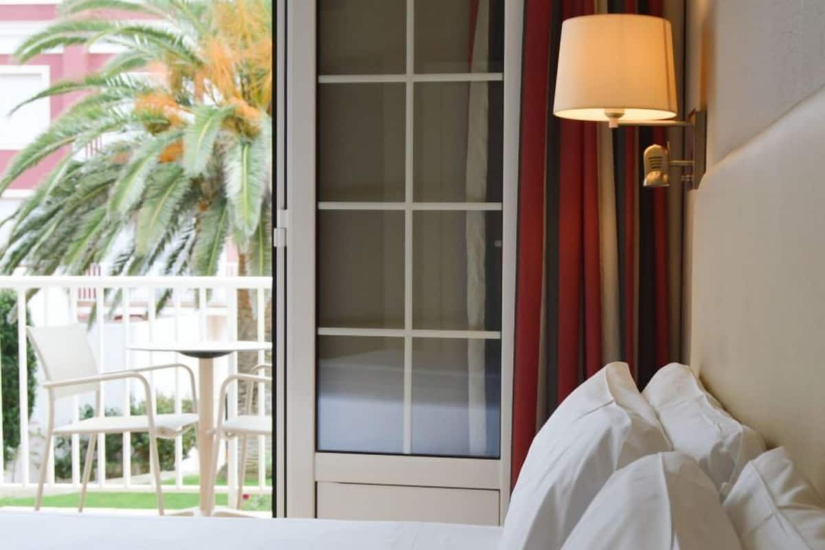 A hotel room with a bed, a window with curtains, a table with chairs, and a palm tree outside.