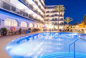 Una piscina con un área hundida, rodeada de mobiliario exterior y un edificio de varios pisos con balcones.