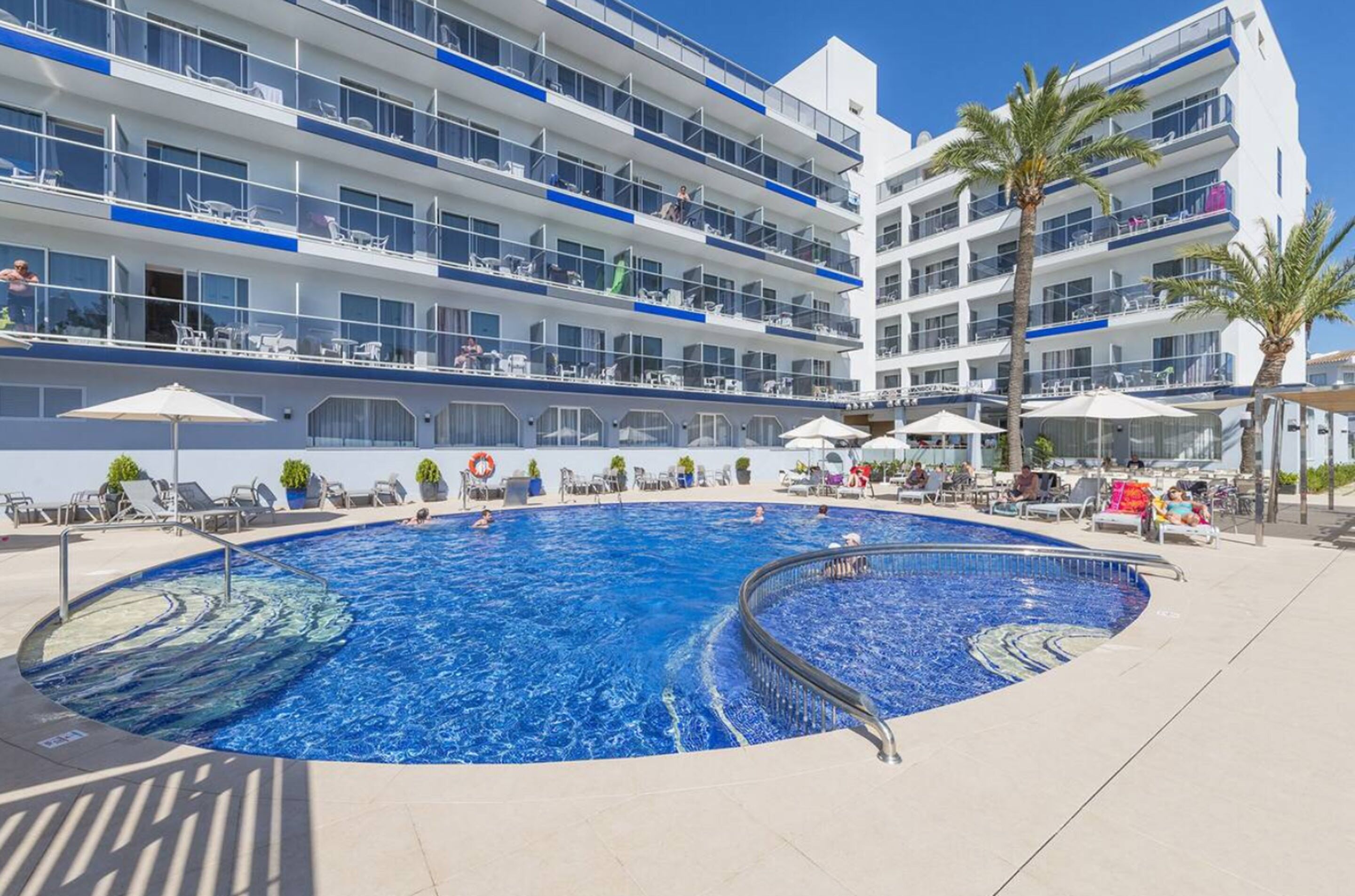 Una piscina con sillas de descanso y sombrillas, rodeada por un edificio de hotel de varios pisos con balcones.
