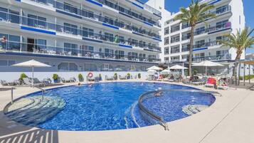 Una piscina con sillas de descanso y sombrillas, rodeada por un edificio de hotel de varios pisos con balcones.