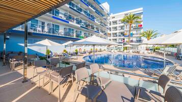 Een zwembadgedeelte van een hotel met meerdere ligbedden en parasols, een groot zwembad en een meerlaags gebouw met balkons.