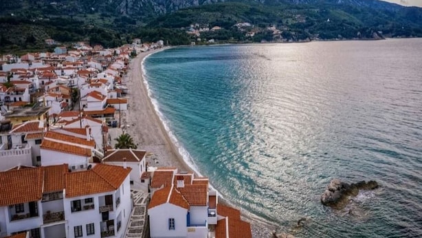 A coastal town with terracotta-roofed buildings along the beach, a clear view of the sea, and mountains in the background. - Blue Sea (Samos)
