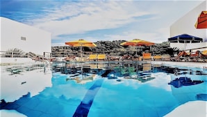 A swimming pool with lounge chairs and umbrellas, surrounded by a white building and greenery. - Blue Sea (Samos)