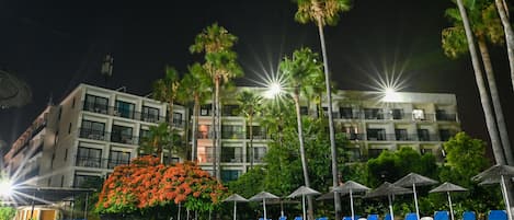 A nighttime pool area with lounge chairs, palm trees, and a building with balconies in the background.