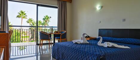 A hotel room with a balcony, a bed with a blue quilted bedspread, two swan-shaped towels, a desk with a chair, and a view of palm trees and buildings.