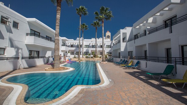 A swimming pool with palm trees, lounge chairs, and a building with balconies. - Anthea Hotel Apartments (Ayia Napa)