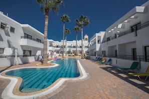 Une piscine bordée de palmiers, avec des chaises longues et un bâtiment doté de balcons.