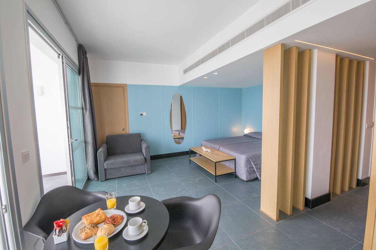 A modern hotel room with a dining area, a sofa, a bed, and a mirror.