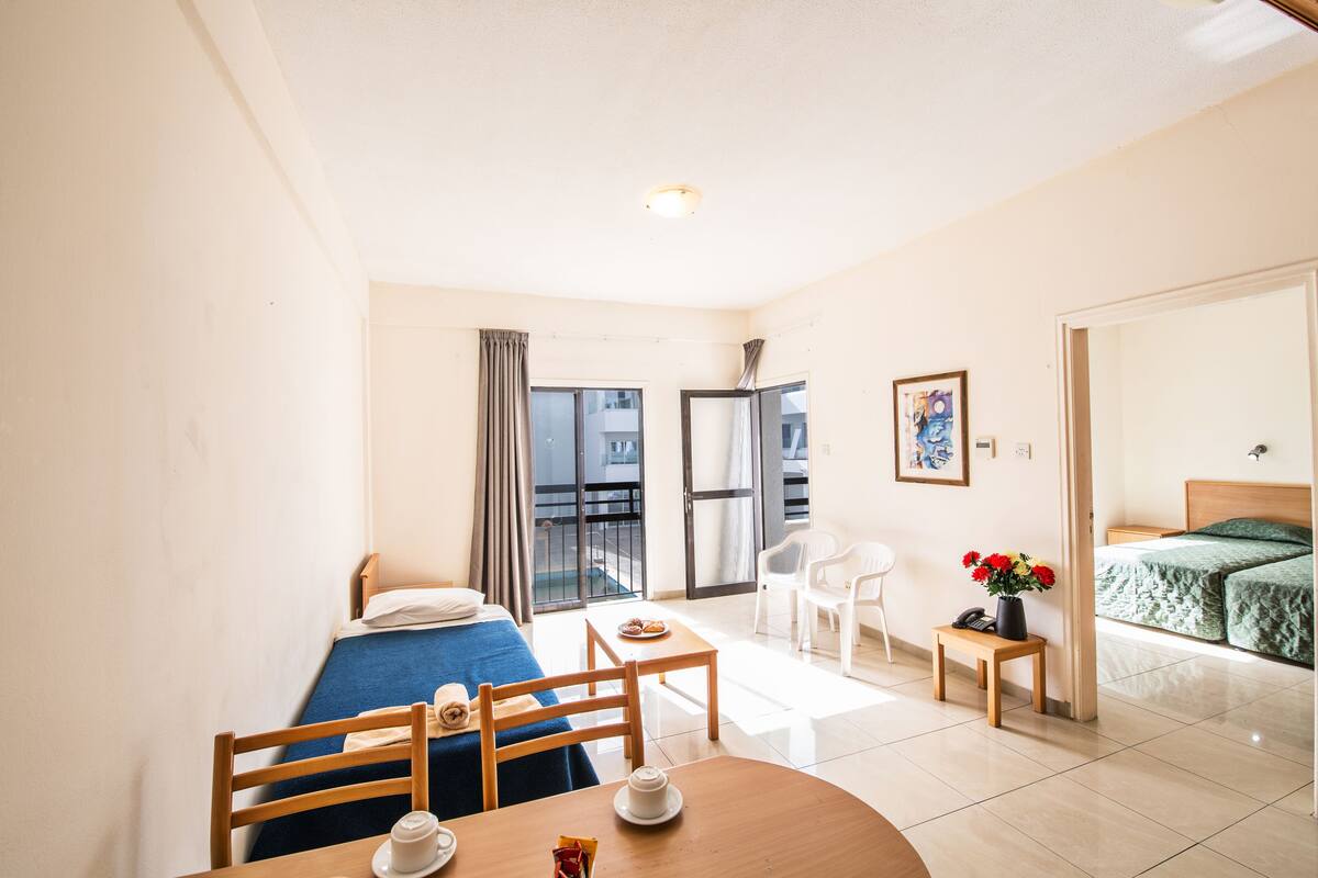 A hotel room with a bed, a small table with a cup and saucer, a chair, a nightstand with a lamp, and a view of the outside through a sliding door.