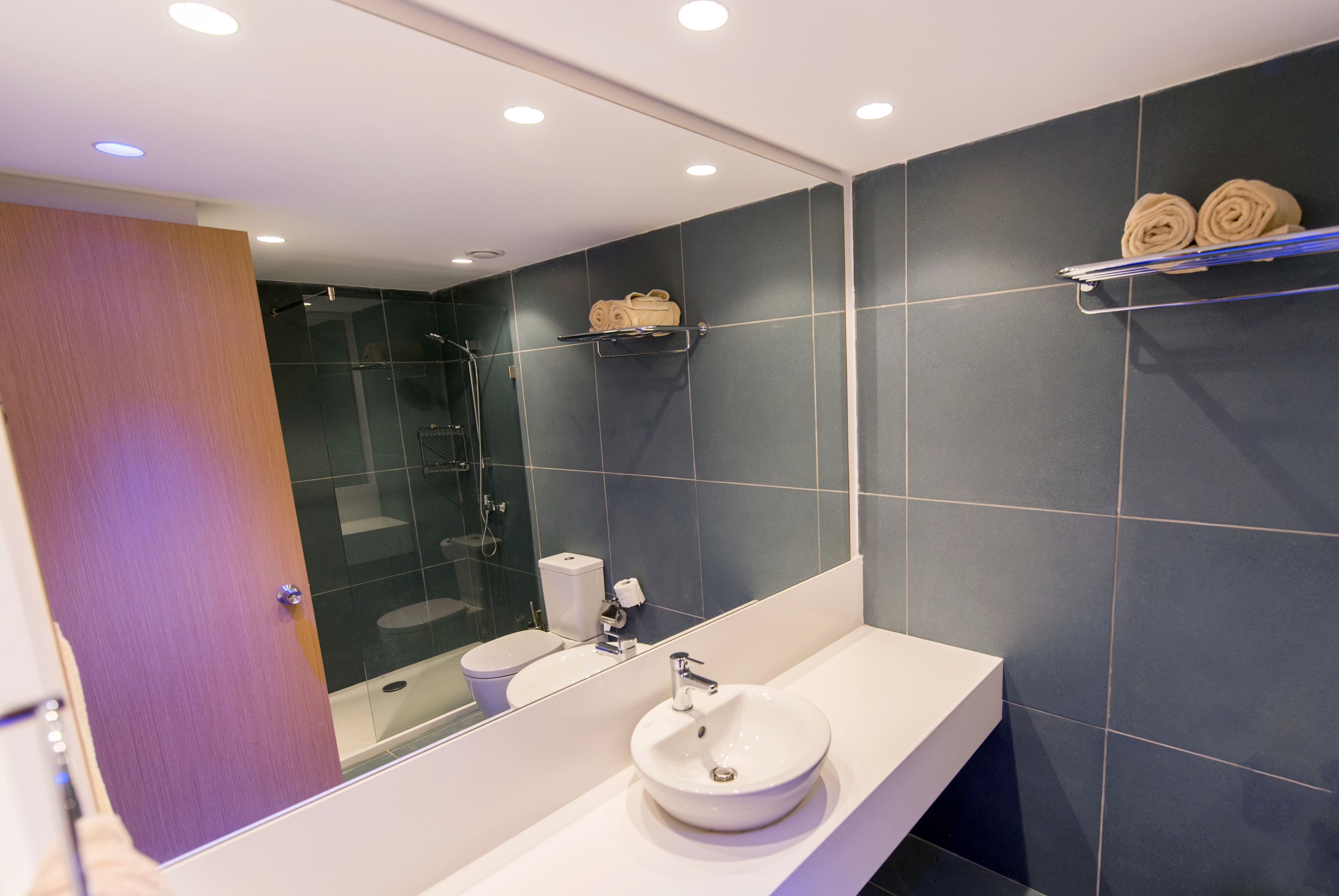 A modern bathroom with a white sink, dark blue tiles, a glass shower enclosure, and towels on a shelf.