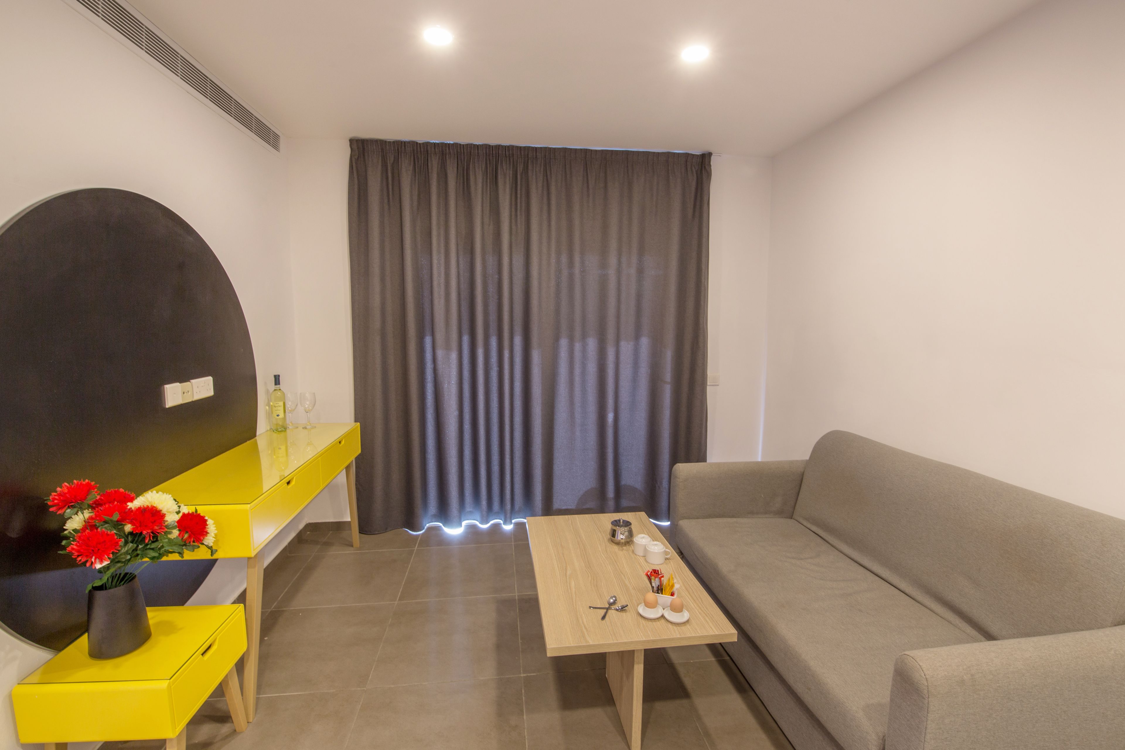 Uma sala de estar moderna com um sofá, uma mesa de centro, uma mesinha lateral amarela com um vaso de flores e um espelho grande na parede.