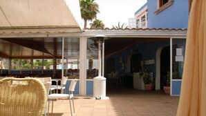 A covered outdoor seating area with chairs and tables, a trash bin, and a building with blue walls and a tiled roof. - Bungalows Las Vegas Golf (San Bartolome de Tirajana)