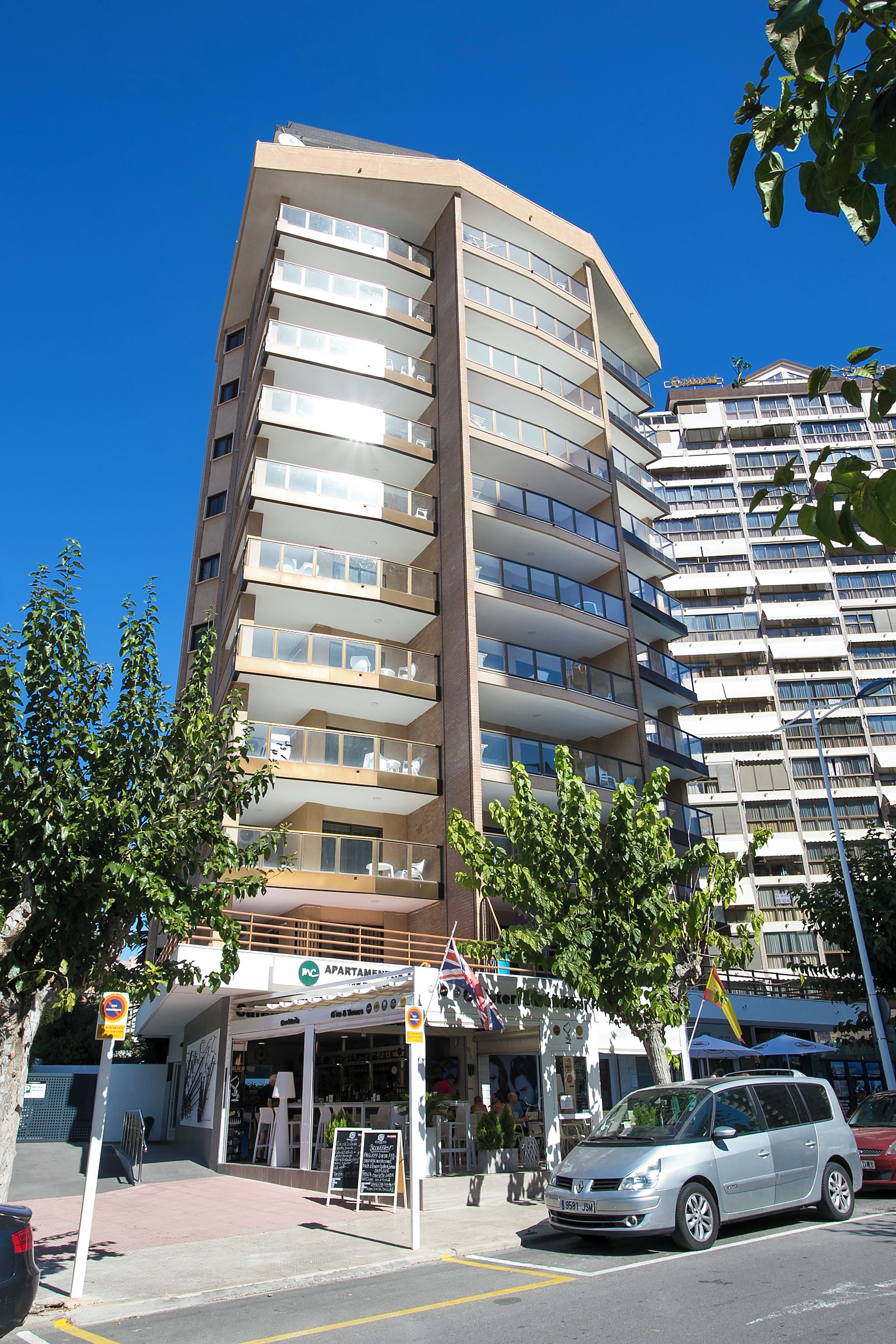 Un edificio de varios pisos con balcones, un café en la planta baja y un estacionamiento.