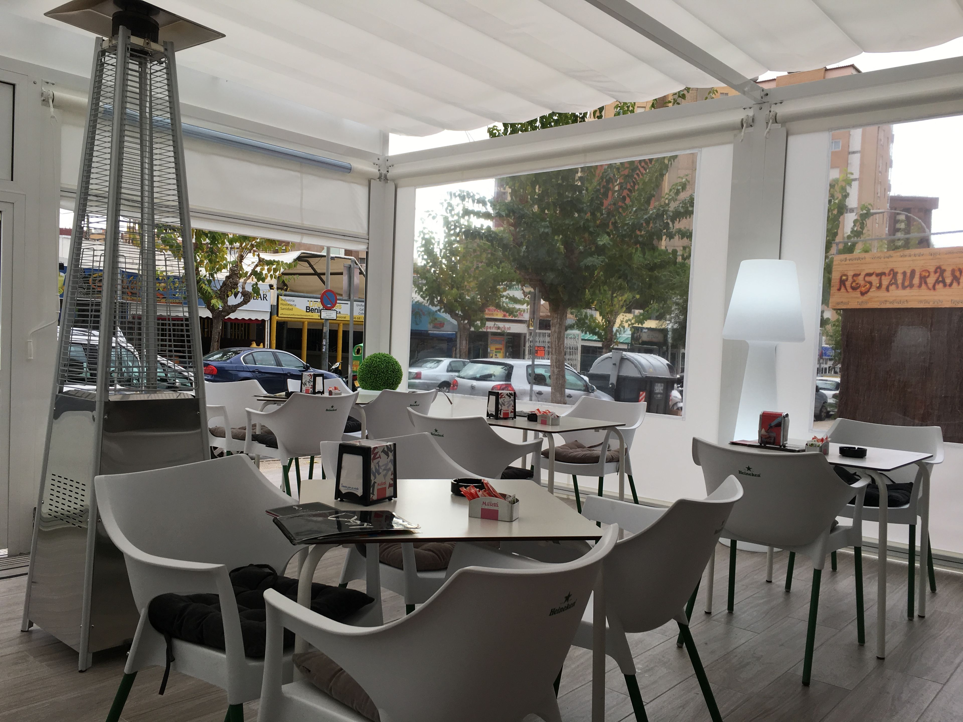 Un área de estar al aire libre con sillas y mesas de plástico blanco, un colgante de luz y vista a la calle con coches estacionados y el letrero de un restaurante.