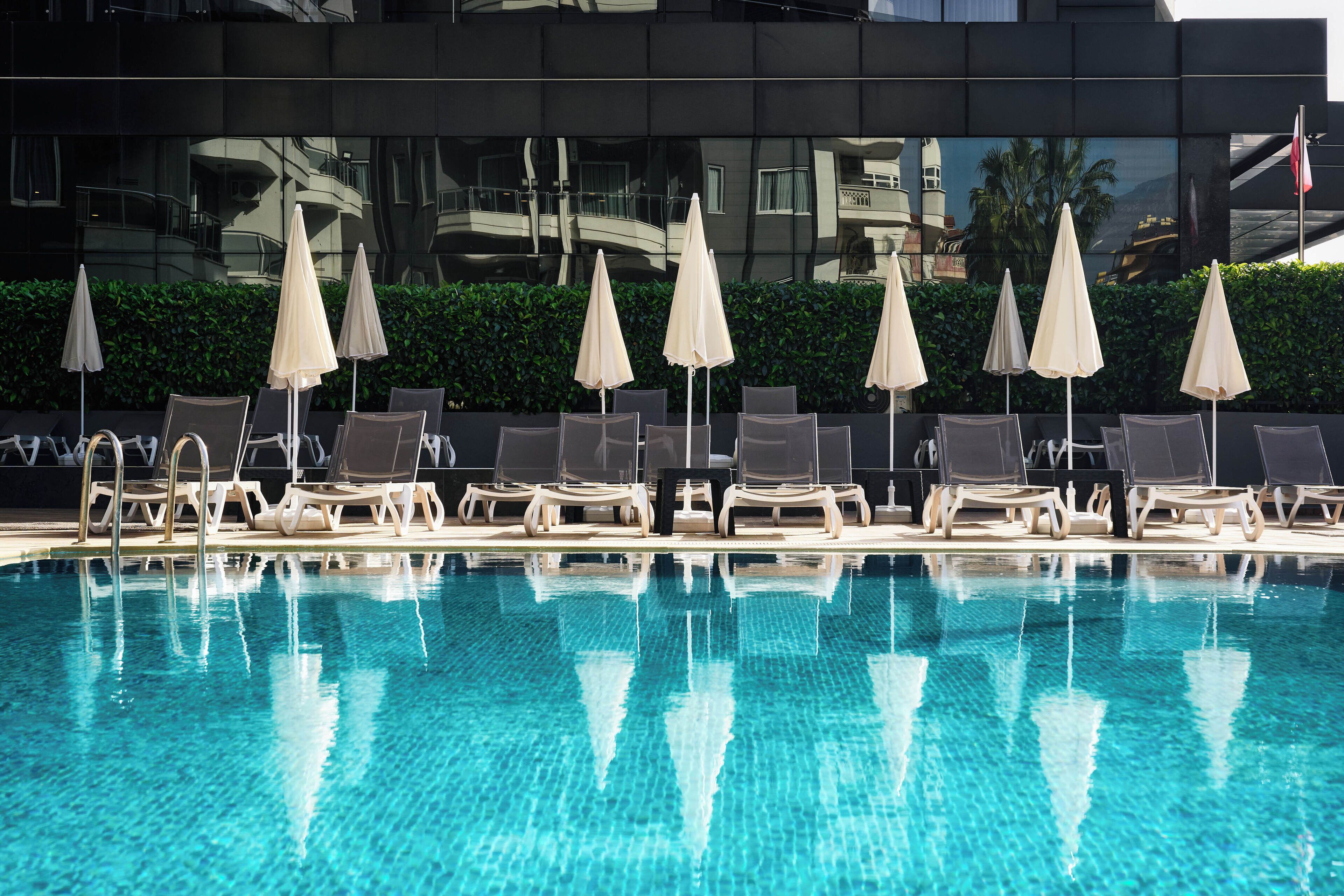 A swimming pool with lounge chairs and umbrellas, reflecting a modern building in the background.