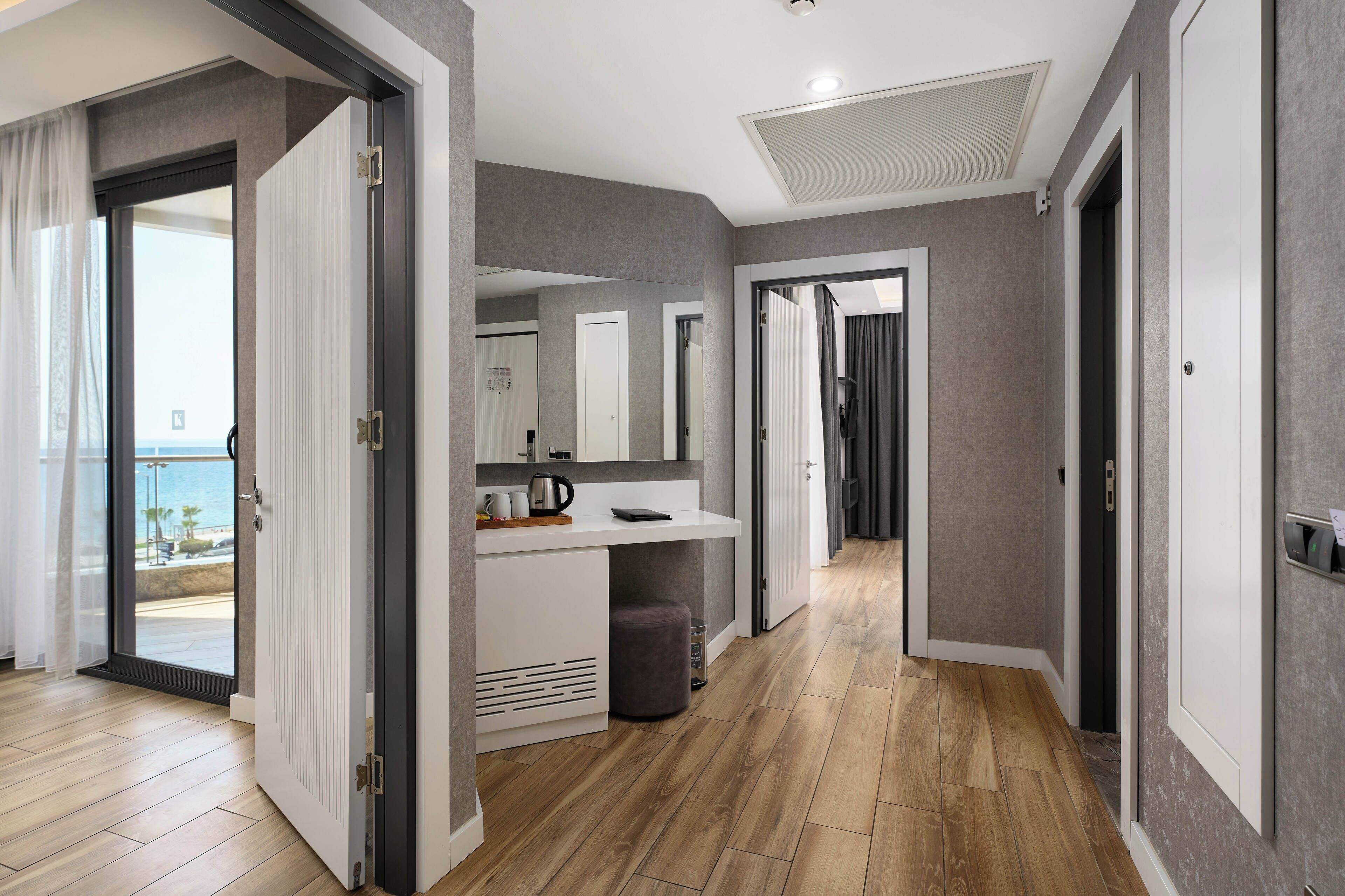 A modern hotel room with a wooden floor, a built-in desk with a mirror, and a view of the sea through a sliding door.