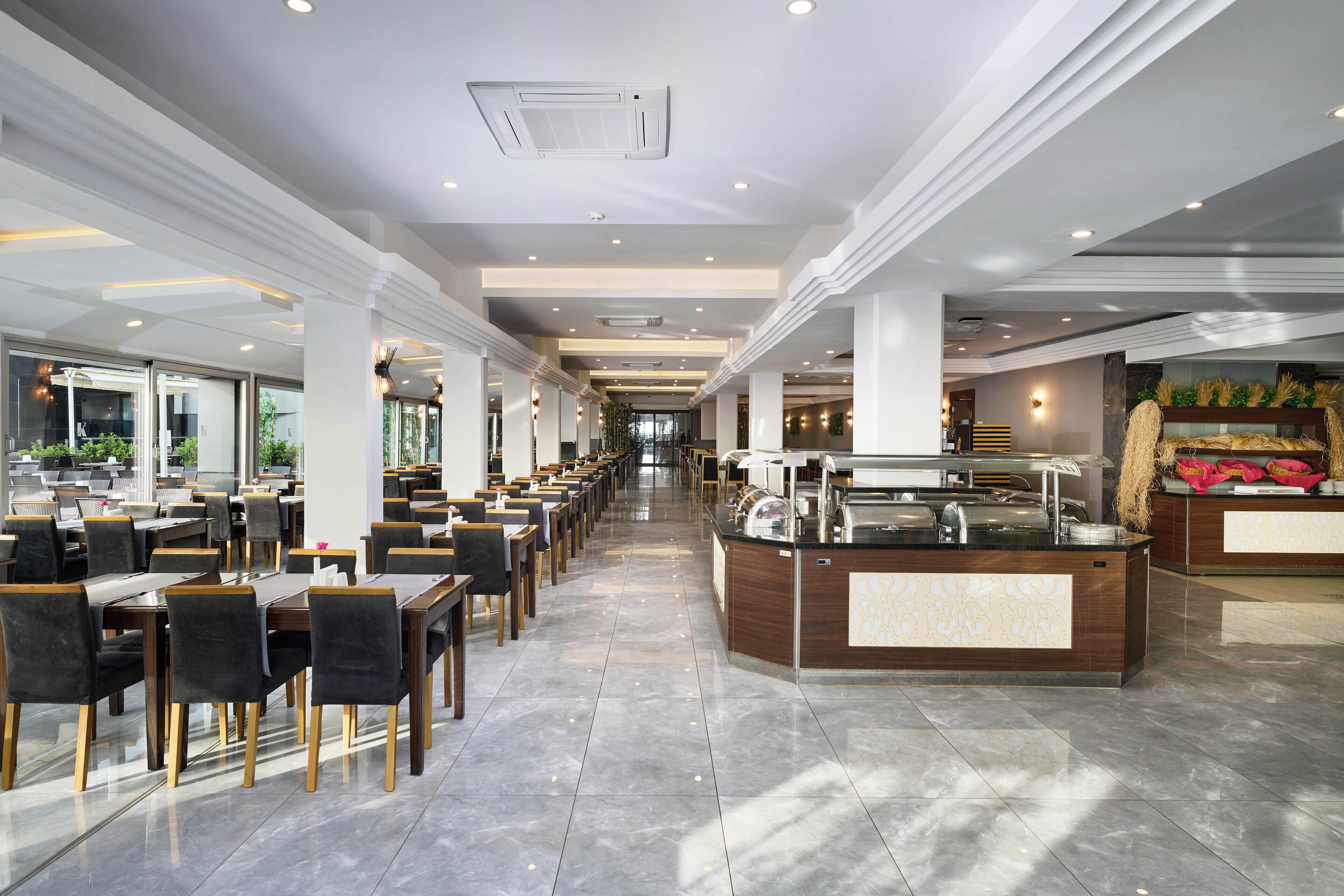 Ein geräumiger Hotel-Speisesaal mit mehreren Tischen und Stühlen, einer zentralen Buffetstation und großen Fenstern, die natürliches Licht hereinlassen.