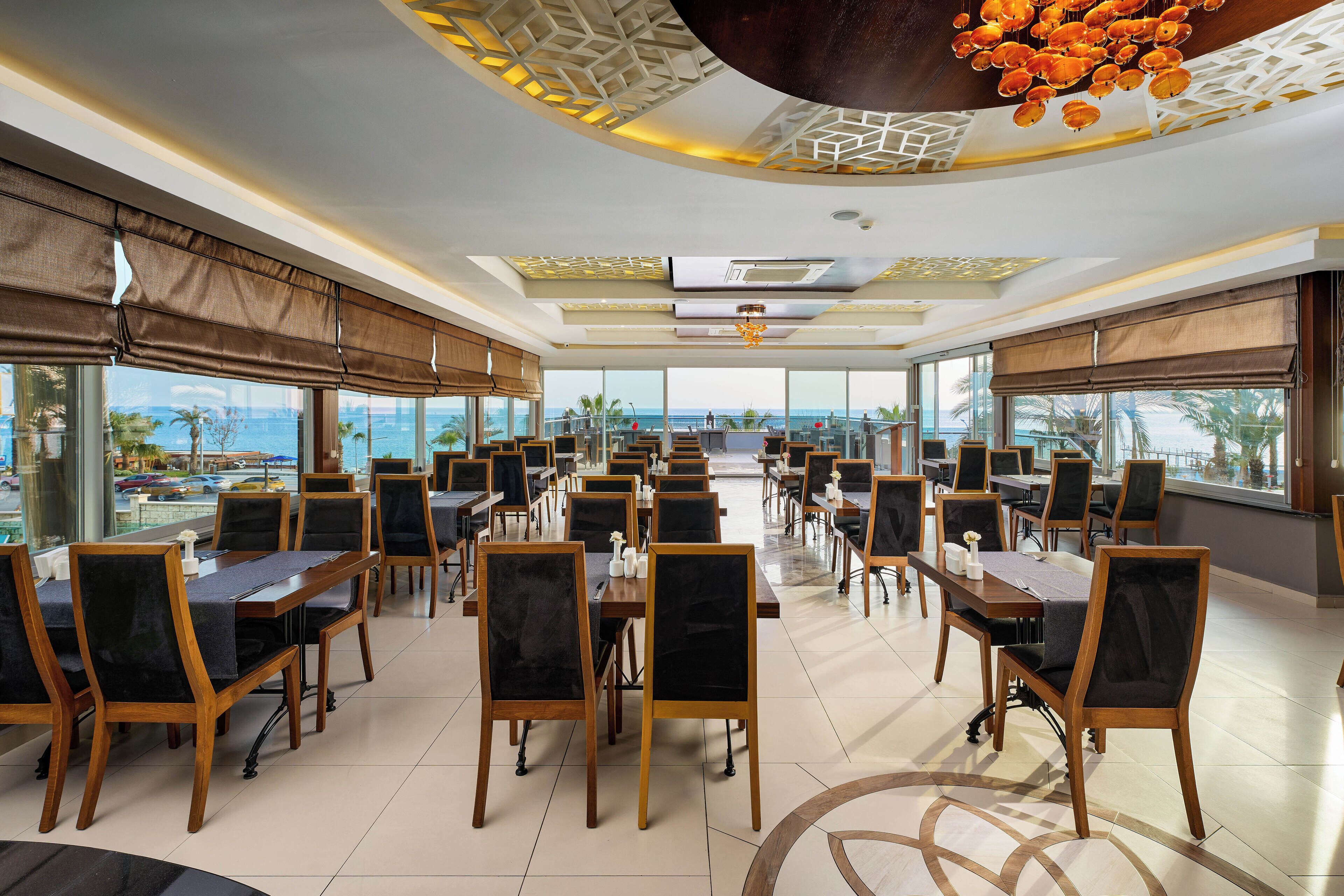A spacious dining area with multiple tables and chairs, large windows offering a view of the sea, and a modern ceiling design.