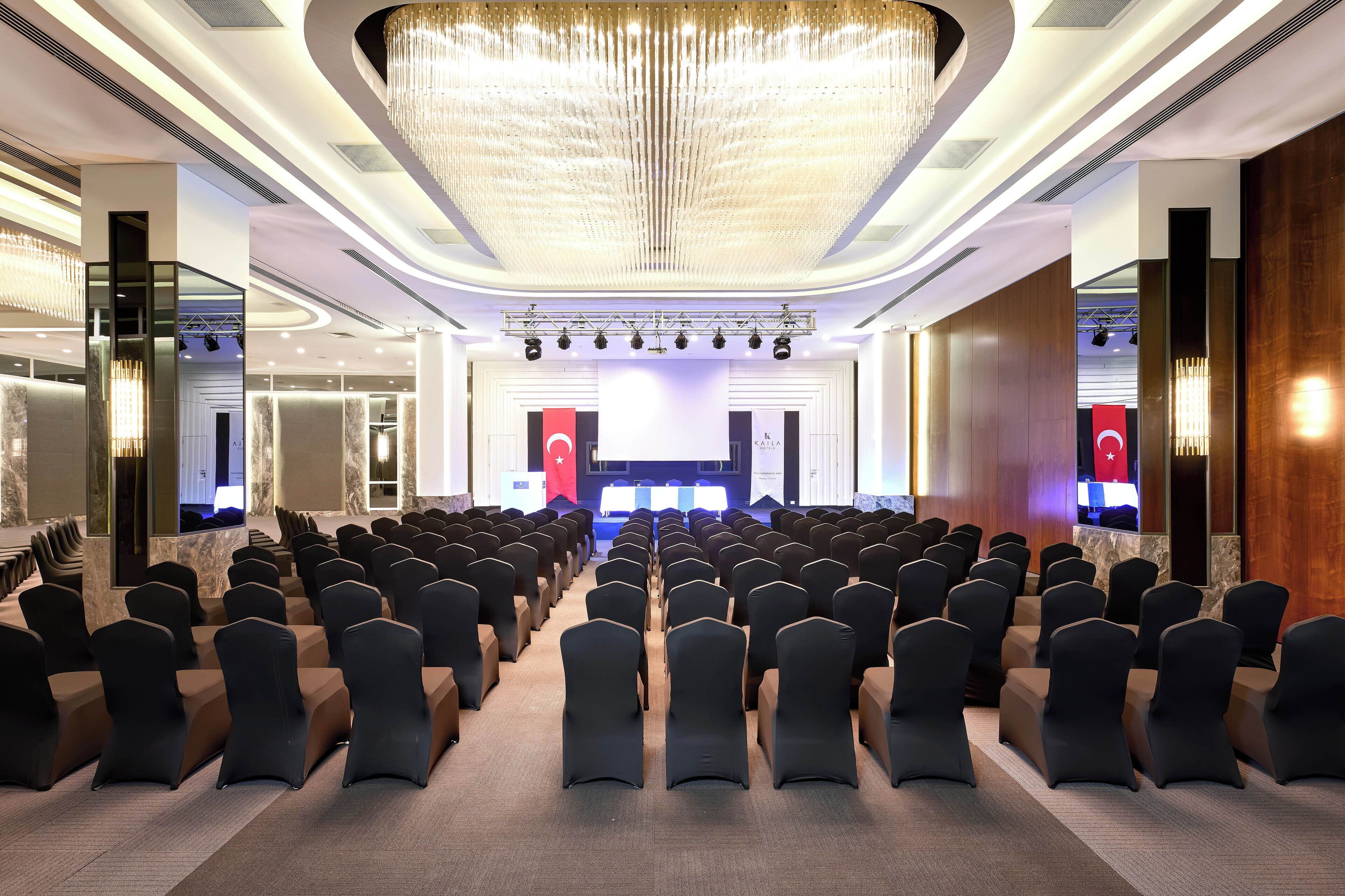 A conference room with rows of chairs, a stage with a podium, and a large chandelier.