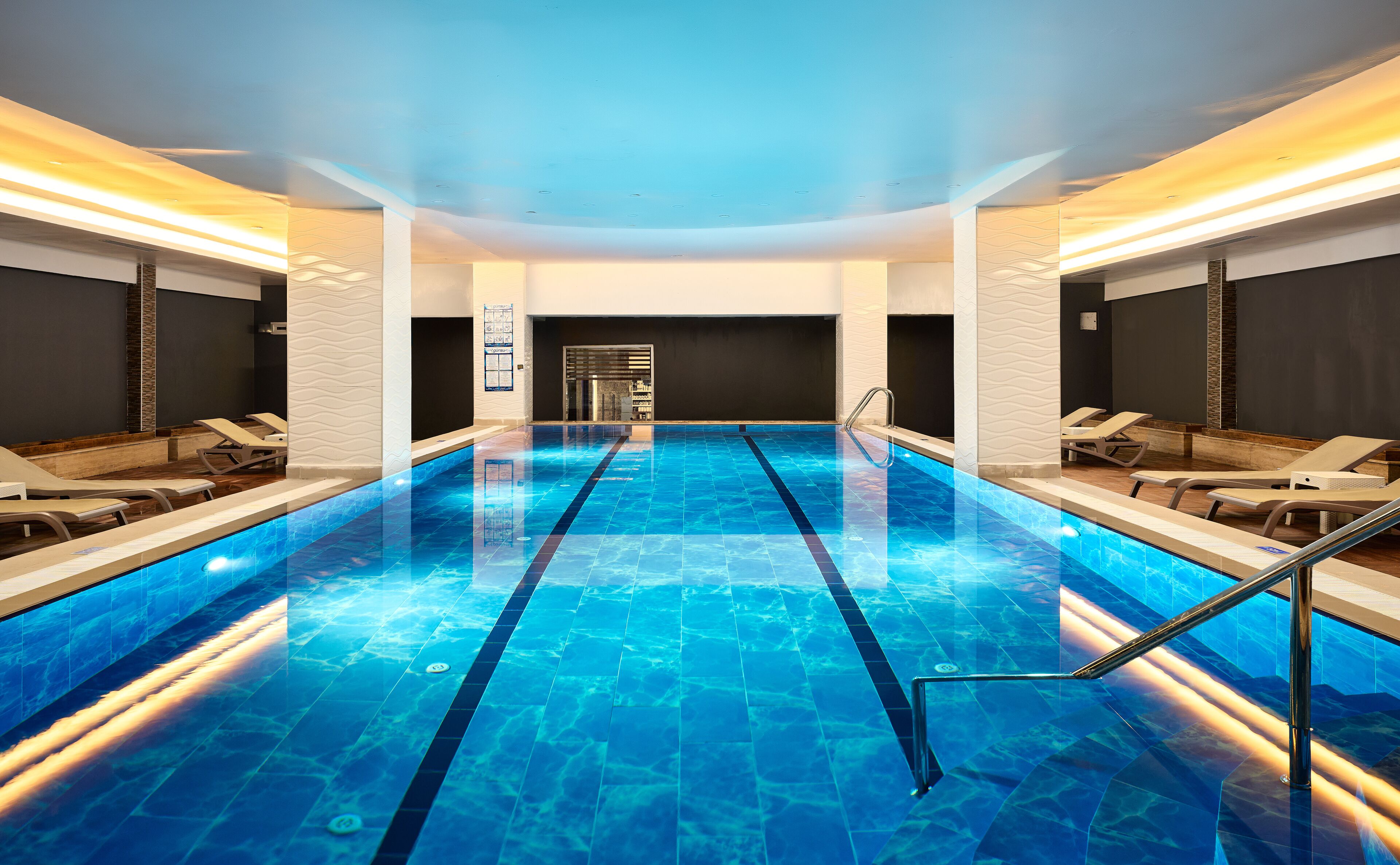 Una piscina interior moderna con baldosas azules brillantes, sillas de descanso y un paseo central.