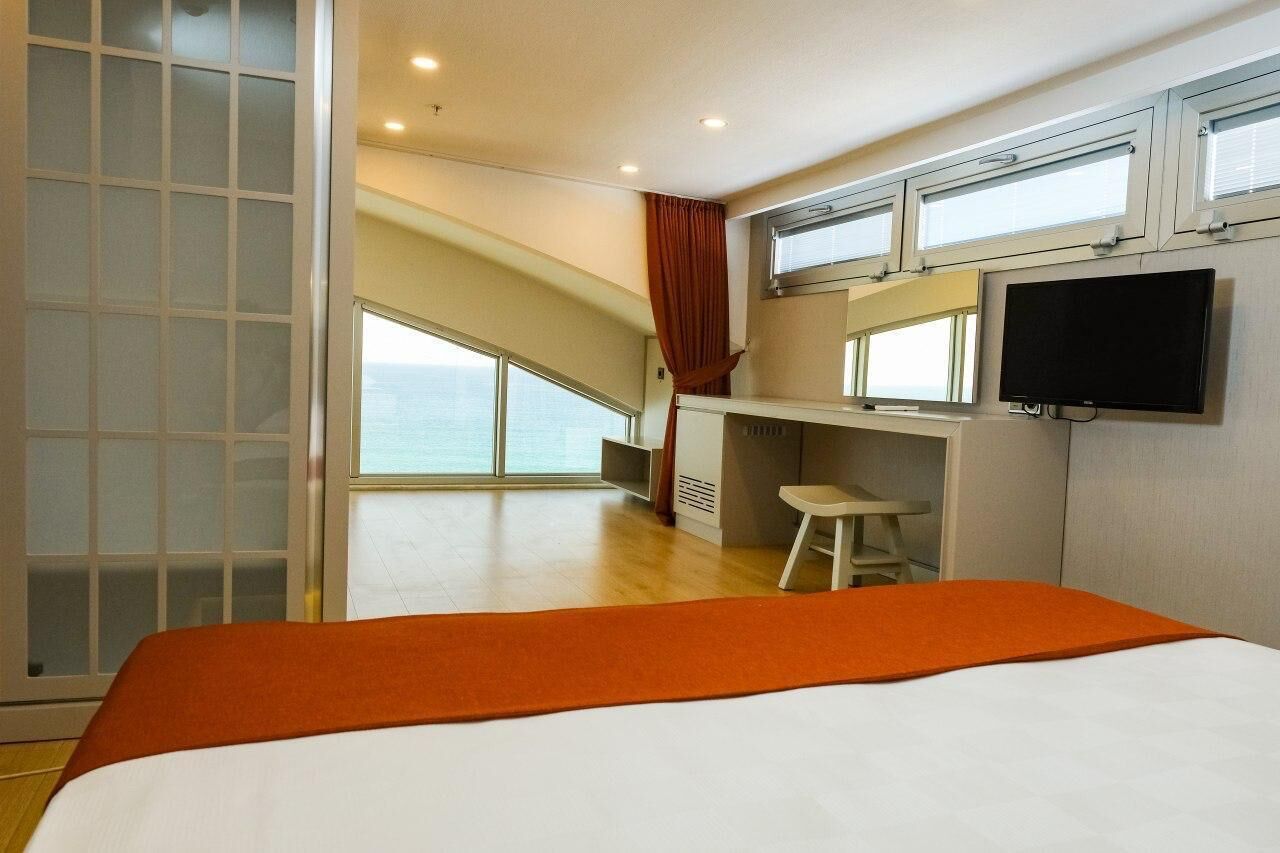 Habitación de hotel con cama, escritorio, televisor y un amplio ventanal con vista al mar.