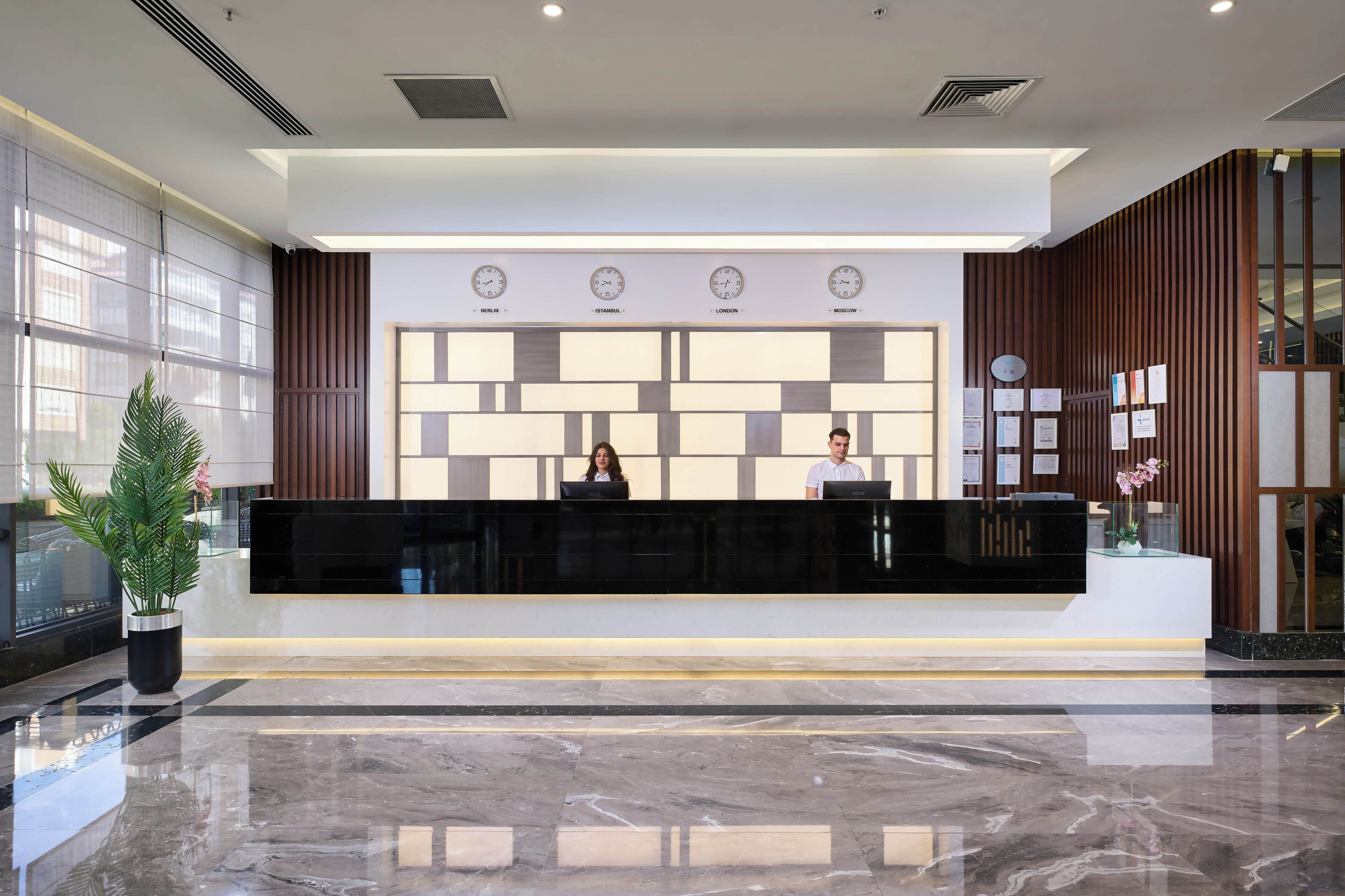 A modern reception desk with digital clocks displaying time in multiple cities, a potted plant, and a clean, polished marble floor.
