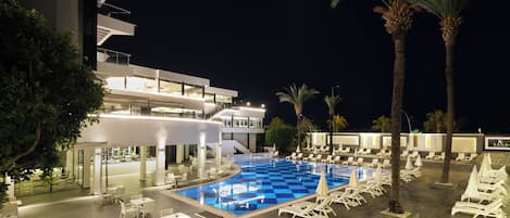 A poolside area with lounge chairs and tables, a swimming pool, and a modern building with balconies.
