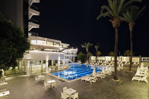Een zwembadterras met ligbedden en tafels, een zwembad en een modern gebouw met balkons.
