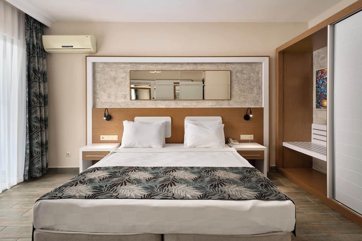 A hotel room with a large bed, a bedside table, a nightstand, a mirror, and an air conditioning unit.