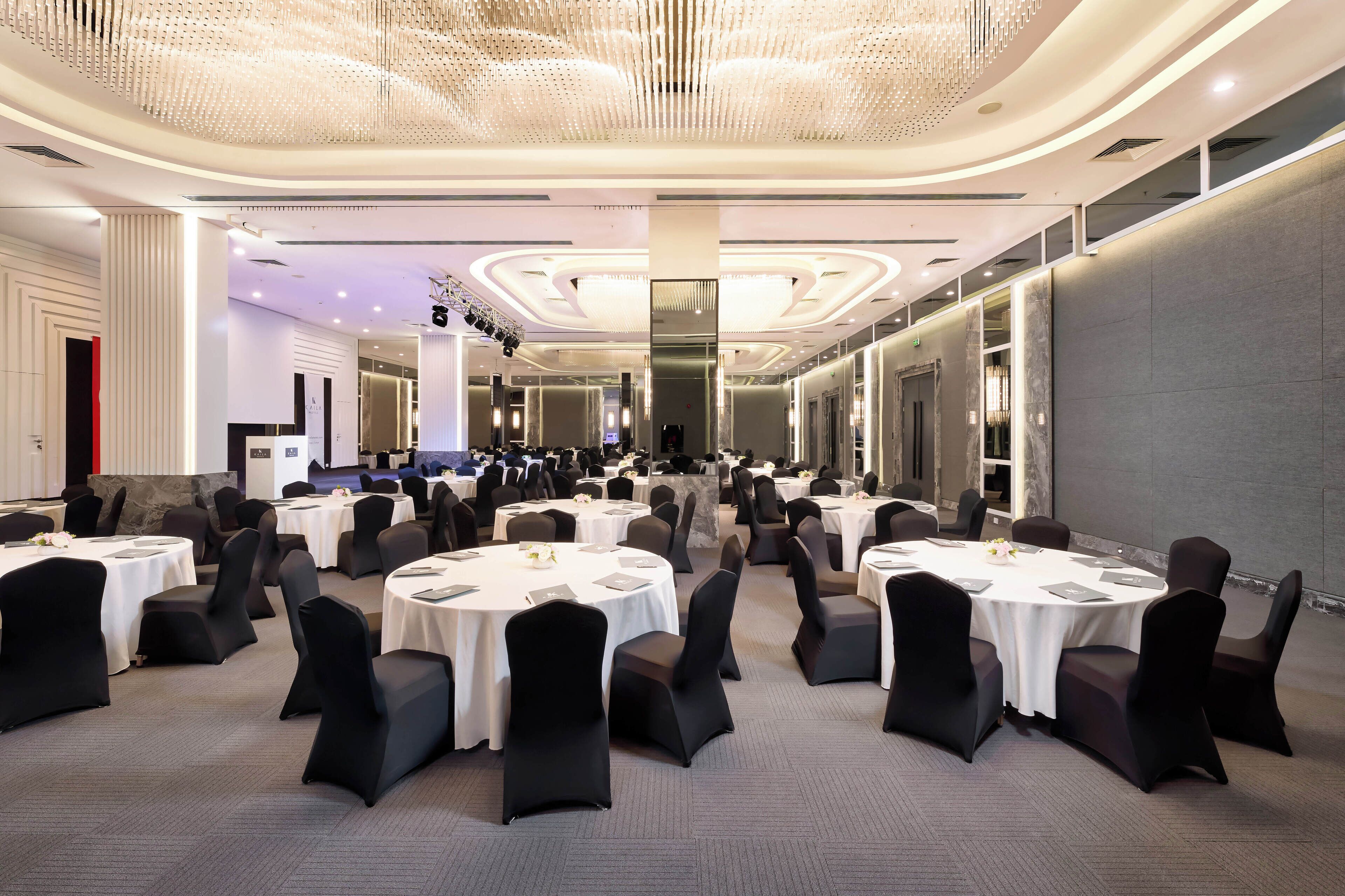 A spacious banquet hall with multiple round tables, black chairs, and a modern chandelier.