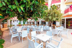 Café al aire libre con mesas y sillas blancas, rodeado de follaje y edificios.